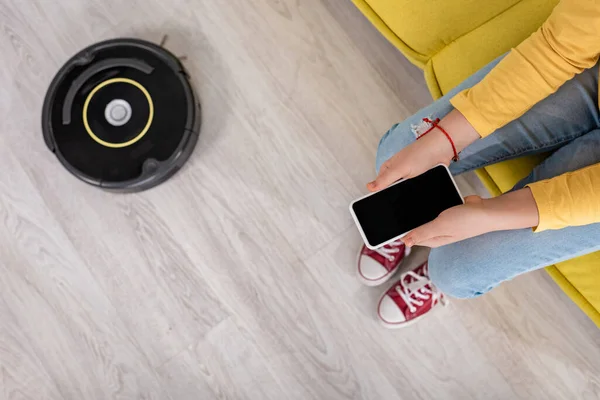 Cropped view of child with smartphone on sofa near robotic vacuum cleaner on floor in living room — Stock Photo