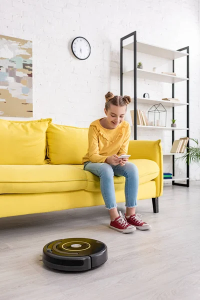 Enfant mignon avec smartphone souriant sur le canapé près de l'aspirateur robotique sur le sol dans le salon — Photo de stock