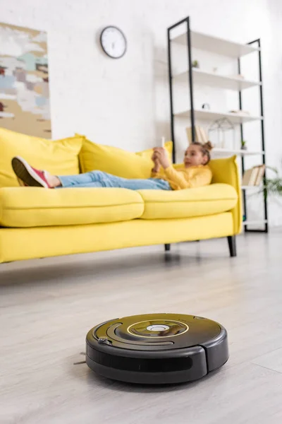 Selective focus of child lying on sofa near robotic vacuum cleaner on floor in living room — Stock Photo