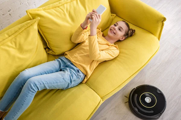 Vista de ángulo alto del niño con teléfono inteligente sonriendo y acostado en el sofá cerca de la aspiradora robótica en el suelo en la sala de estar - foto de stock