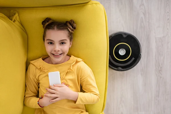 Vue du dessus de mignon enfant avec smartphone souriant, regardant la caméra et couché sur le canapé près de l'aspirateur robotique sur le sol dans le salon — Photo de stock