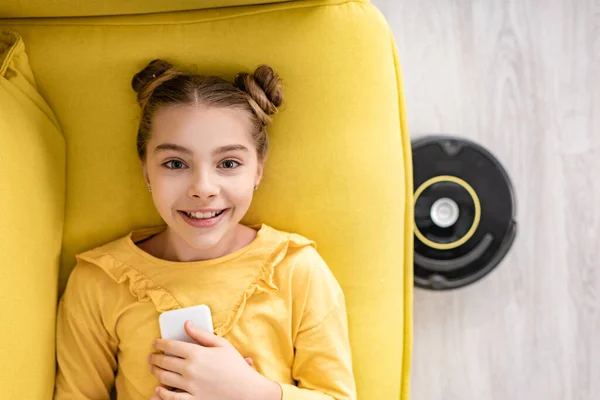 Vue du dessus de mignon enfant avec smartphone souriant, regardant la caméra et couché sur le canapé près de l'aspirateur robotique sur le sol dans le salon — Photo de stock