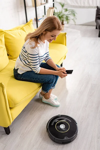 Vista de ángulo alto de la mujer sonriendo con el teléfono inteligente en el sofá cerca de la aspiradora robótica en el suelo en la sala de estar - foto de stock