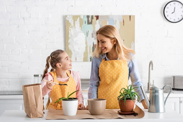 Mãe e filha olhando umas para as outras e sorrindo perto da mesa com panela de rega, saco de papel e vasos de flores na cozinha — Fotografia de Stock