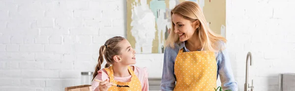 Orientação panorâmica de mãe e filha olhando uma para a outra e sorrindo na cozinha — Fotografia de Stock