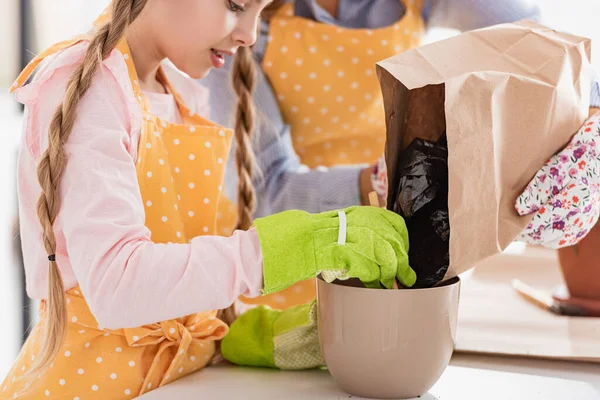 Vista ritagliata di donna mettendo a terra vaso da fiori con bambino concentrato con pala vicino tavolo in cucina — Foto stock