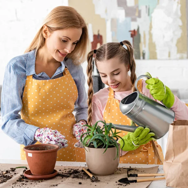 Mother and cute daughter watering aloe in flowerpot near gardening tools on table in kitchen — Stock Photo