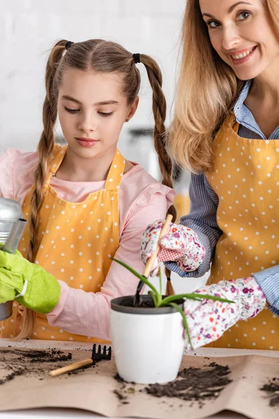 Woman with shovel and flowerpot with aloe smiling and cute daughter holding watering pot near table in kitchen — Stock Photo