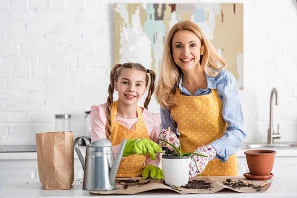 Madre y linda hija sonriendo con herramientas de jardinería y maceta con aloe cerca de la olla de riego y bolsa de papel con tierra en la mesa - foto de stock