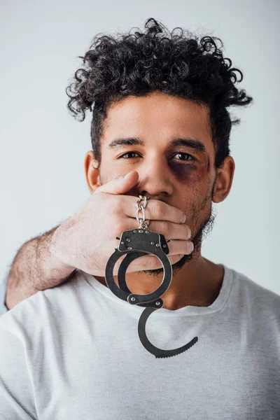 Male hand holding handcuffs and covering mouth to african american man with bruise isolated on grey — Stock Photo