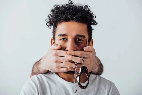 Male hands with handcuffs covering mouth to african american man with bruise isolated on grey — Stock Photo