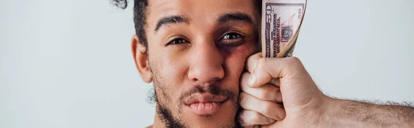 Мужская рука держит долларовые банкноты у лица африканского американца с синяком, изолированным на сером, панорамном снимке — стоковое фото