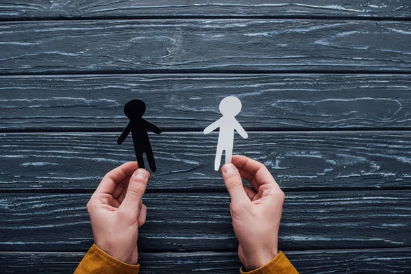 Top view of man holding black and white human figures on dark wooden background — Stock Photo