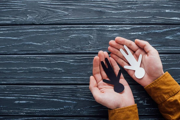 Vista superior de papel figuras humanas preto e branco em mãos masculinas sobre fundo de madeira escura — Fotografia de Stock