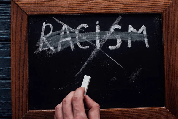 Top view of man holding chalk near blackboard with racism lettering on dark wooden background — Stock Photo
