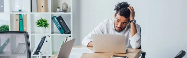 Cultura panorâmica do empresário afro-americano usando laptop perto de papéis e smartphones na mesa no escritório — Fotografia de Stock