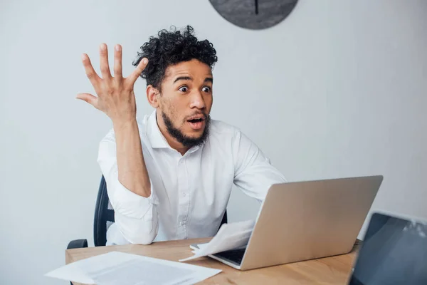 Selektiver Fokus eines schockierten afrikanisch-amerikanischen Geschäftsmannes, der im Büro neben Laptops und Dokumenten auf dem Tisch sitzt — Stockfoto