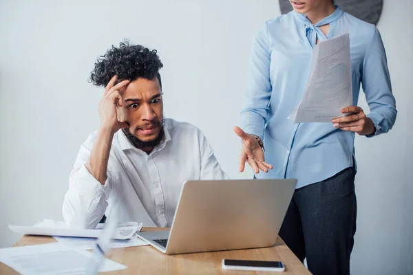 Selective focus of businesswoman holding papers and pointing at laptop to scared african american man in office — Stock Photo