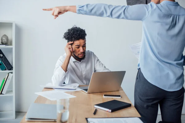 Selektiver Fokus eines afrikanisch-amerikanischen Mannes mit Laptop in der Nähe einer Geschäftsfrau, die im Büro mit dem Finger zeigt — Stockfoto