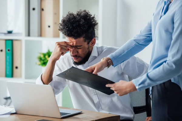 Selective focus of businesswoman holding clipboard near sad african american businessman at table — Stock Photo