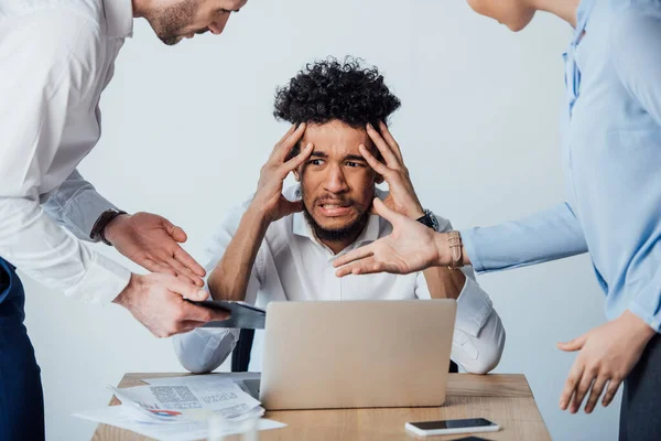 Enfoque selectivo de la gente de negocios hablando en el hombre afroamericano asustado cerca de documentos y gadgets en la mesa - foto de stock