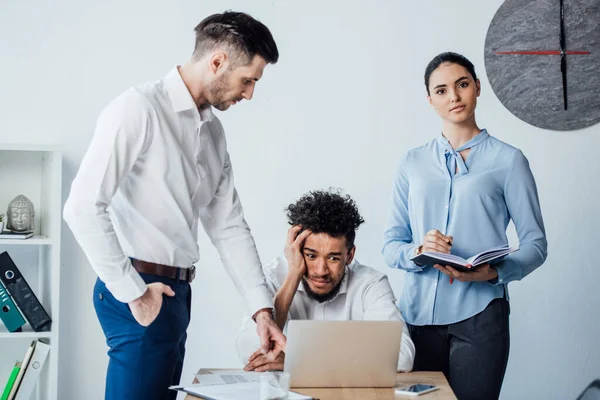 Des hommes d'affaires multiethniques se tenant près d'un homme d'affaires afro-américain confus à table au bureau — Photo de stock