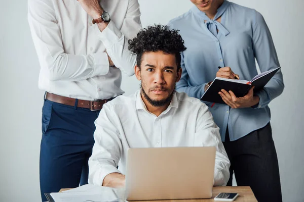 Enfoque selectivo del hombre de negocios afroamericano mirando a la cámara cerca de gente de negocios con portátil en la oficina - foto de stock