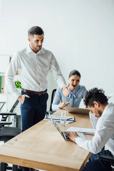 Selective focus of multicultural business people pointing at african american businessman using laptop n office — Stock Photo