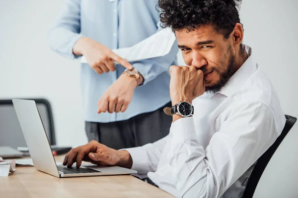 Вибірковий фокус афроамериканського бізнесмена, який дивиться на камеру під час роботи над ноутбуком біля колеги, що вказує на наручний годинник. — стокове фото