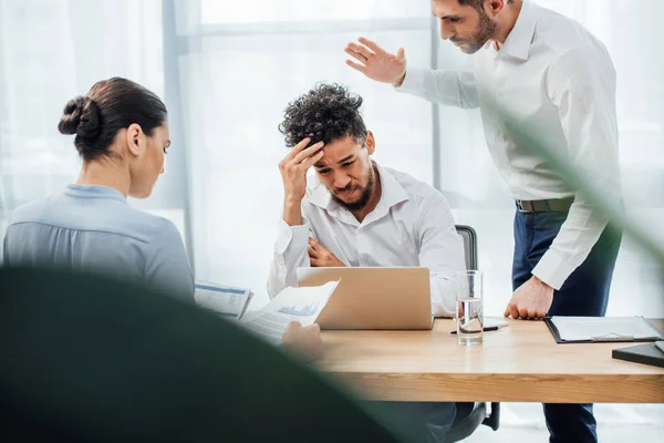 Focus selettivo dell'uomo d'affari che litiga con un triste collega afroamericano vicino a laptop e documenti o tavolo — Foto stock