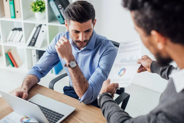 Selective focus of african american businessman holding paper with charts near colleague in office — Stock Photo