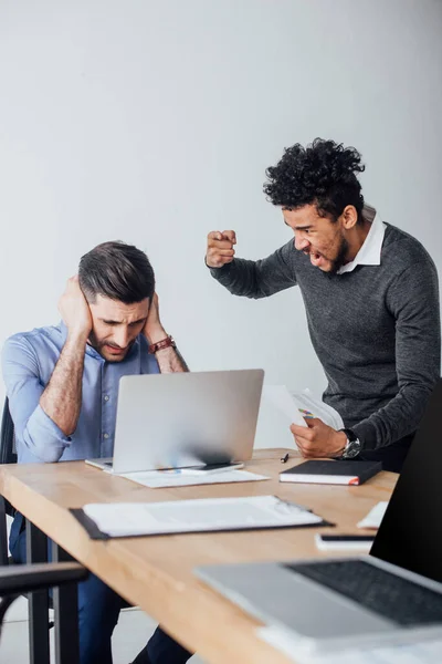 Selective focus of angry african american businessman quarreling at colleague covering ears in office — Stock Photo