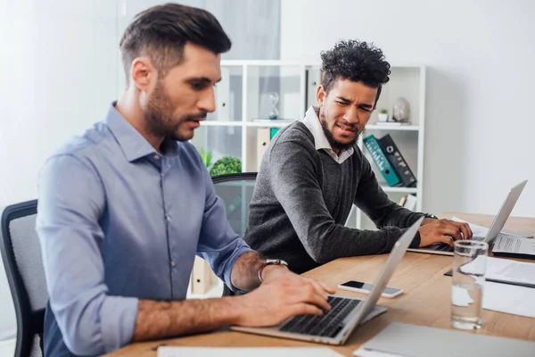 Focus selettivo di confuso uomo d'affari afro-americano guardando collega che utilizza il computer portatile in ufficio — Foto stock