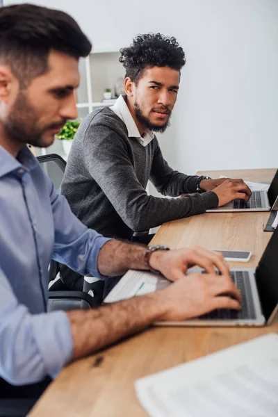 Selective focus of skeptical african american businessman looking at colleague using laptop near papers in office — Stock Photo