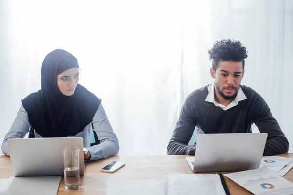 Enfoque selectivo de la mujer de negocios musulmana mirando a un colega afroamericano mientras trabaja en el cargo - foto de stock