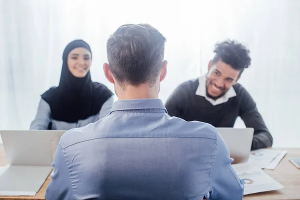 Foco seletivo do empresário sentado perto de colegas multiculturais sorridentes no cargo — Fotografia de Stock