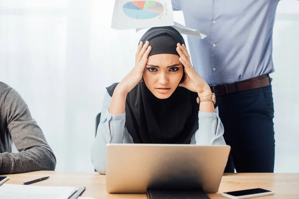 Triste mujer de negocios árabe mirando la cámara cerca de colegas en la oficina - foto de stock