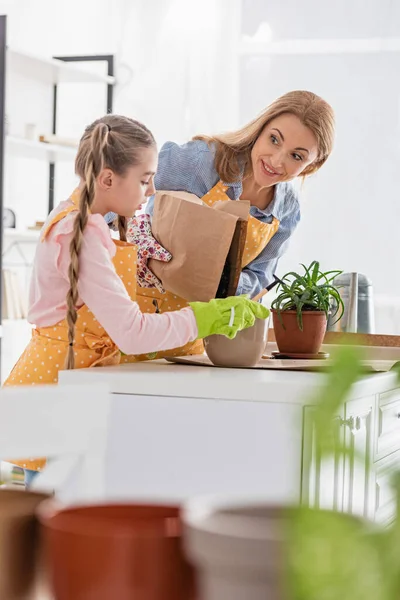 Focus selettivo della madre sorpresa che mette a terra il vaso da fiori e guarda la figlia con rastrello vicino al tavolo in cucina — Foto stock