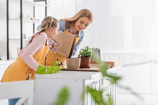 Selektiver Fokus der Mutter lächelnd und Boden für Blumentopf und Tochter mit Gartengeräten am Tisch in der Küche — Stockfoto