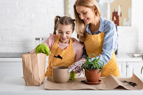 Mãe e filha bonito colocando chão para vaso com pá perto da mesa com saco de papel e aloe na cozinha — Fotografia de Stock