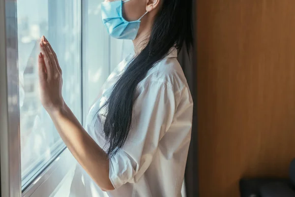 Ausgeschnittene Ansicht einer jungen Frau in medizinischer Maske, die am Fenster ihres Hauses steht — Stockfoto