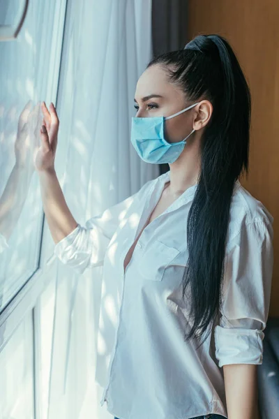 Chica triste en máscara médica mirando a través de la ventana en casa - foto de stock