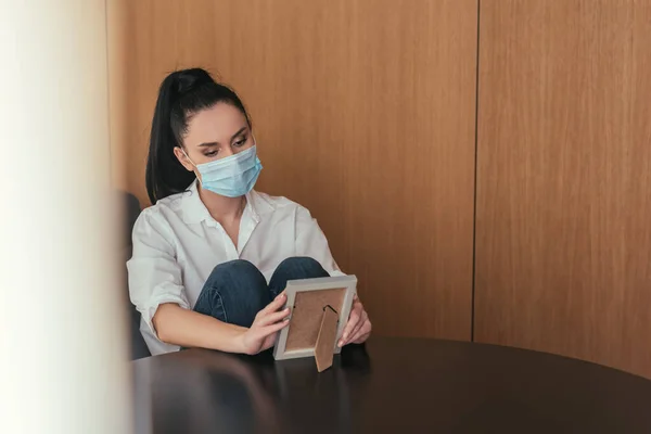Selective focus of sad young woman in protective mask sitting at table and looking at photo frame — Stock Photo