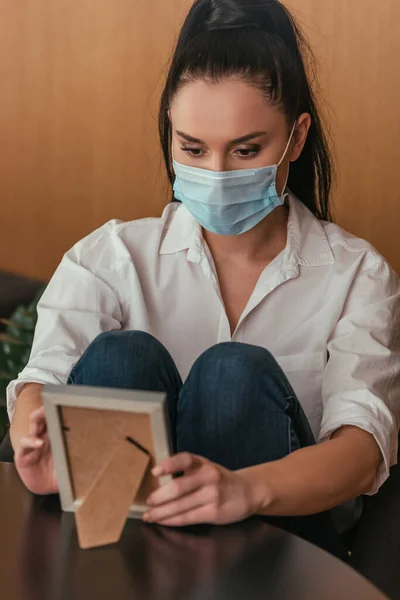 Sad young woman in protective mask sitting at table and looking at photo frame — Stock Photo