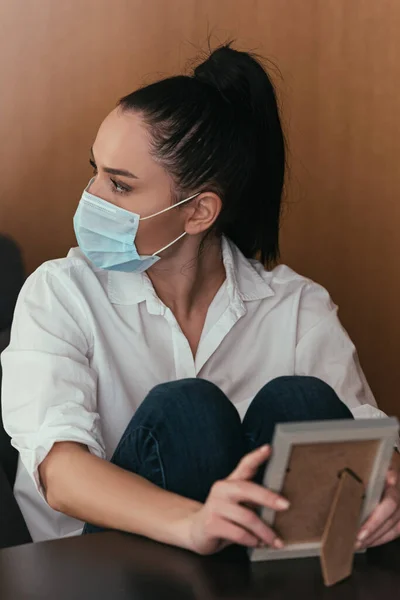 Depressive junge Frau in medizinischer Maske schaut weg, während sie Fotorahmen hält — Stockfoto