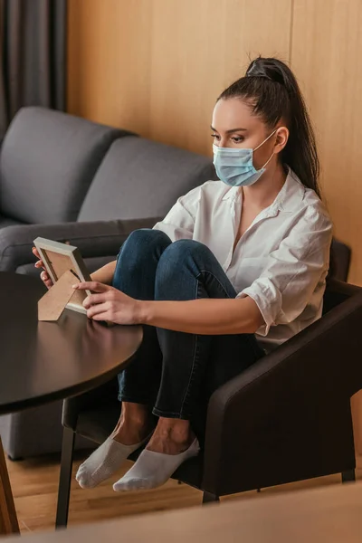 Chica triste en máscara médica mirando marco de la foto mientras está sentado en el sillón cerca de la mesa - foto de stock