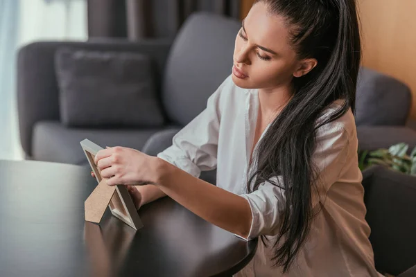 Triste joven mujer mirando marco de fotos mientras está sentado en la mesa - foto de stock