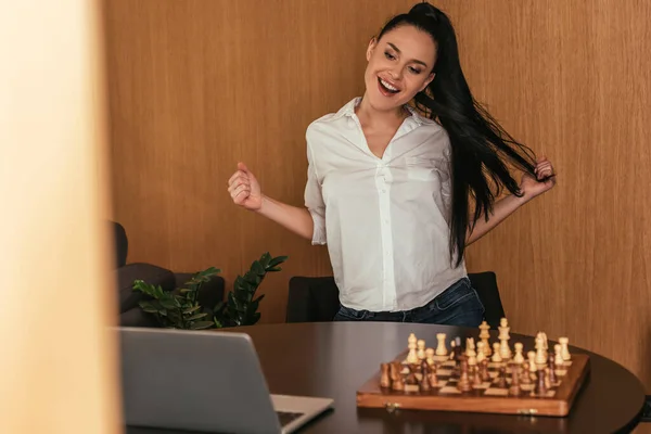 Foyer sélectif de femme excitée montrant geste gagnant près de l'échiquier et ordinateur portable sur la table — Photo de stock