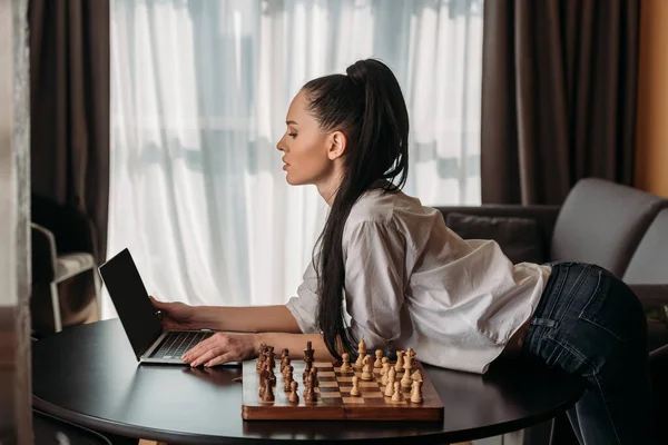 Side view of attractive woman looking at laptop with blank screen near chessboard on table — Stock Photo