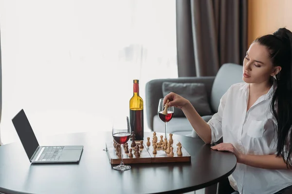 Mulher atraente jogando xadrez perto de laptop com tela em branco e vinho tinto na mesa — Fotografia de Stock